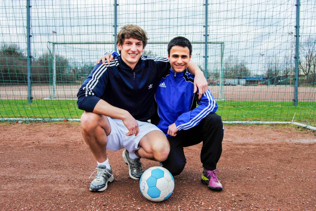Zwei lächelnde Teilnehmer des Mentoring-Programms ROCK YOUR LIFE! knien vor einem Fußballtor und umarmen sich.
